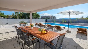 Un espacio exterior techado con piscina, sillas de descanso y una mesa de comedor dispuesta con fruta.