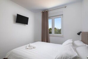 Un dormitorio con cama, televisor montado en la pared, ventana con cortinas y vista a zonas verdes.