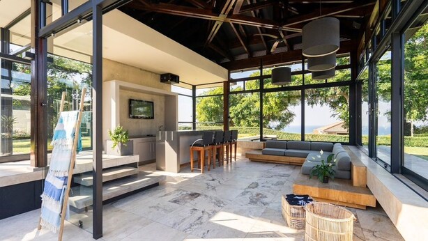 Un espacio exterior moderno para disfrutar, con cocina, asientos y grandes ventanales que dan a un paisaje pintoresco.