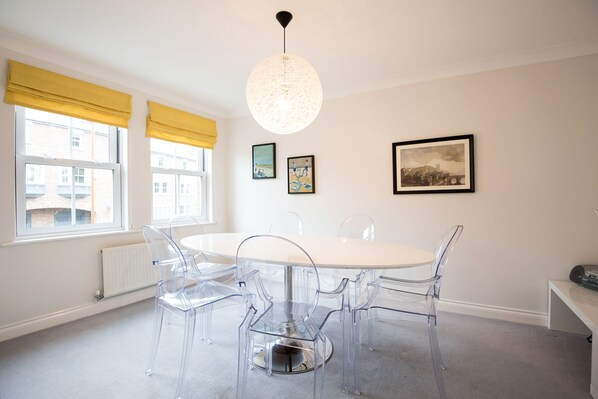 A modern dining area with a round table and clear chairs, large windows with yellow curtains, and artwork on the walls.