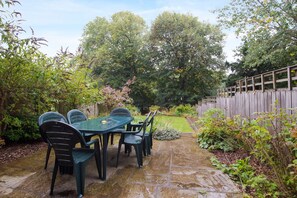A stone patio with a table and chairs, surrounded by lush greenery and a wooden fence. - Dalton Crescent (Durham)