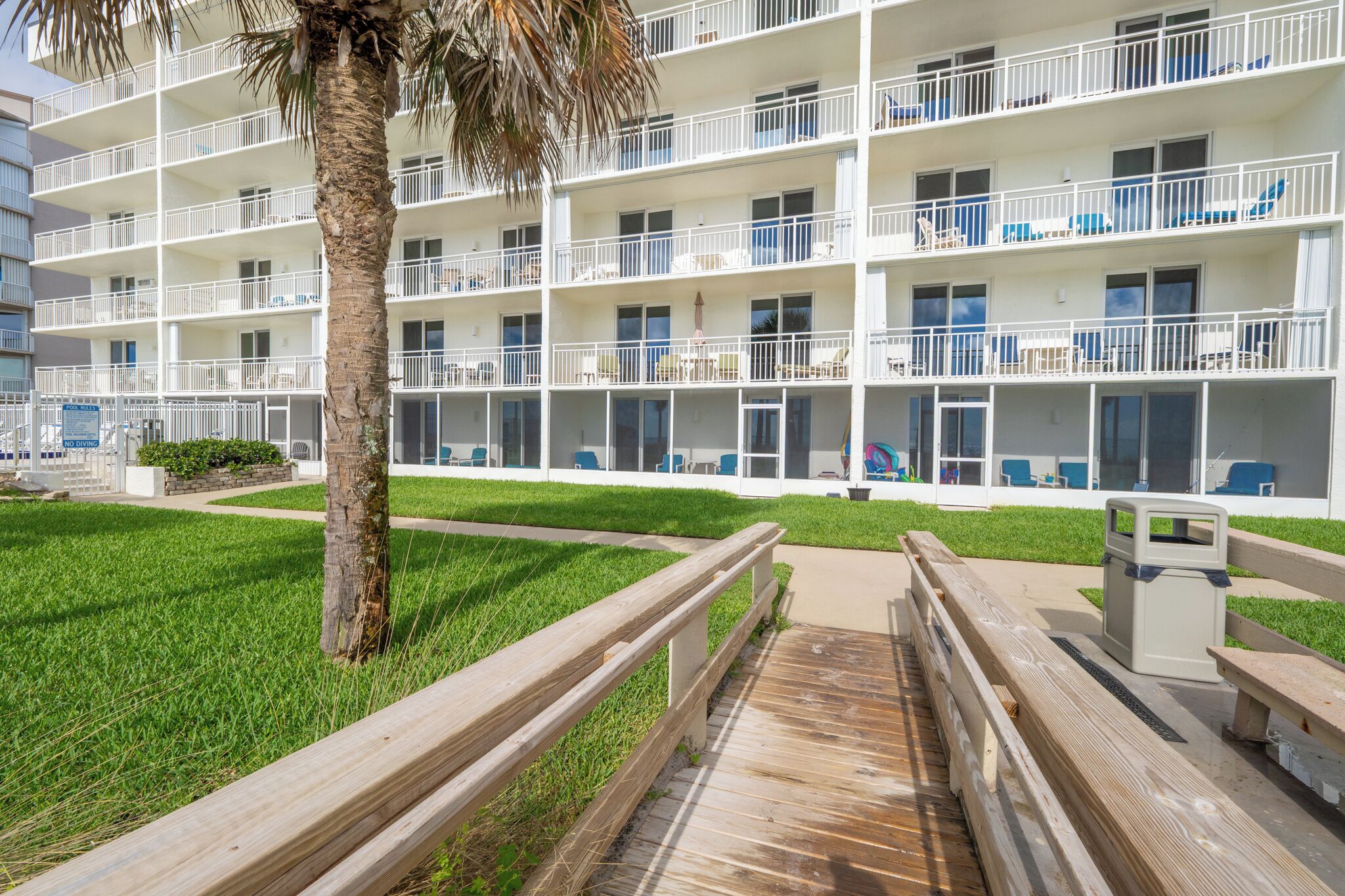A multi-story hotel with balconies, a palm tree, and a wooden boardwalk.