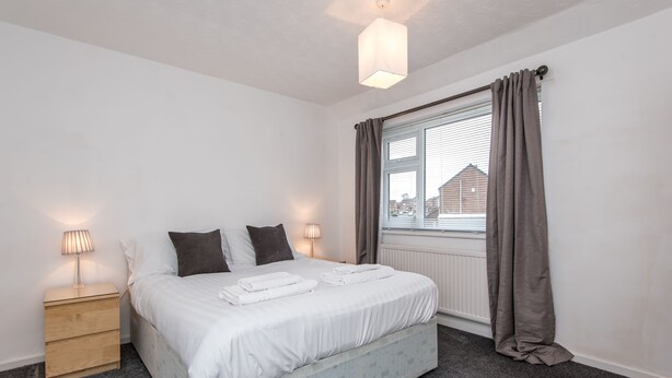 A bedroom with a bed, bedside lamps, a window with curtains, and a view of buildings outside. - Broome Road (Durham)