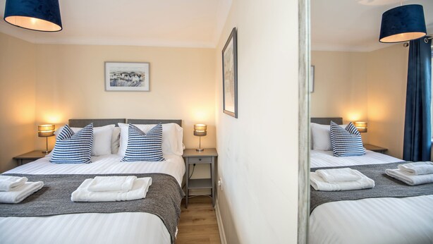 A hotel room with two beds, each with a grey bedspread, white linens, and blue and white checkered pillows. There are two bedside tables with lamps, a framed picture on the wall, and a mirror reflecting the room. - Hillbrook (Stockton-on-Tees)
