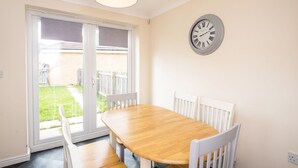 A dining area with a wooden table and white chairs, a wall clock, and a sliding glass door leading to a backyard. - Hillbrook (Stockton-on-Tees)
