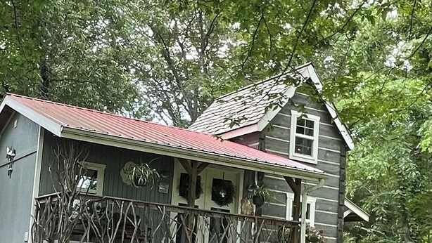 A rustic cabin with a stone foundation, a wooden porch with a railing, and a red roof. - Cedar Twig Cabin: Serene and cozy (Mt. Juliet)