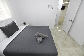 Cama bem arrumada com colcha cinza escura e toalha enrolada sobre ela, um travesseiro preto e uma pequena planta em vaso em uma mesinha de cabeceira.