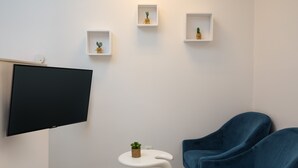 En moderne stue med et fladskærms-tv, et hvidt, buet sofabord, en blå sofa og tre små hylder med planter.