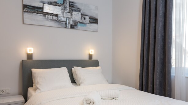 En pænt redt seng med hvide sengetæpper og puder, et natbord med en lampe, og et moderne abstrakt billede på væggen.