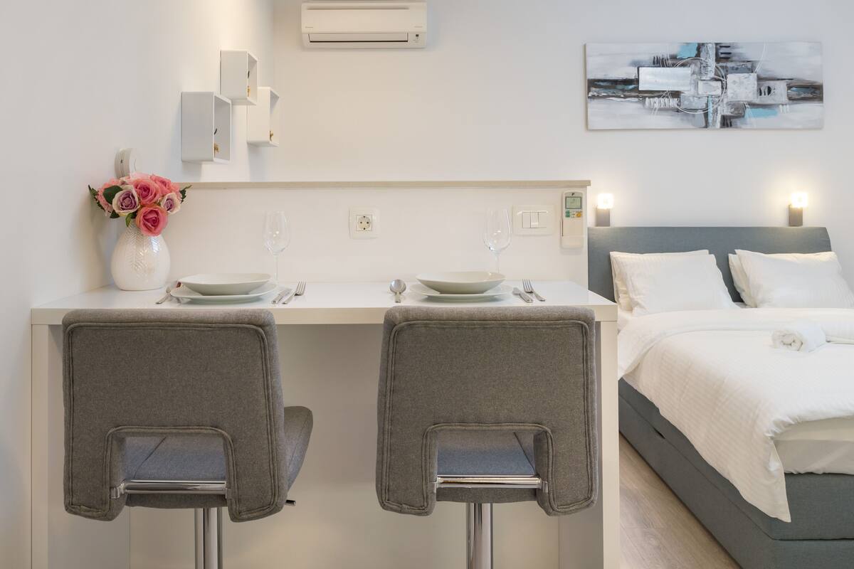Une chambre d’hôtel moderne, équipée d’un lit, d’une petite kitchenette, de deux chaises, d’un vase avec des fleurs et d’un tableau au mur.