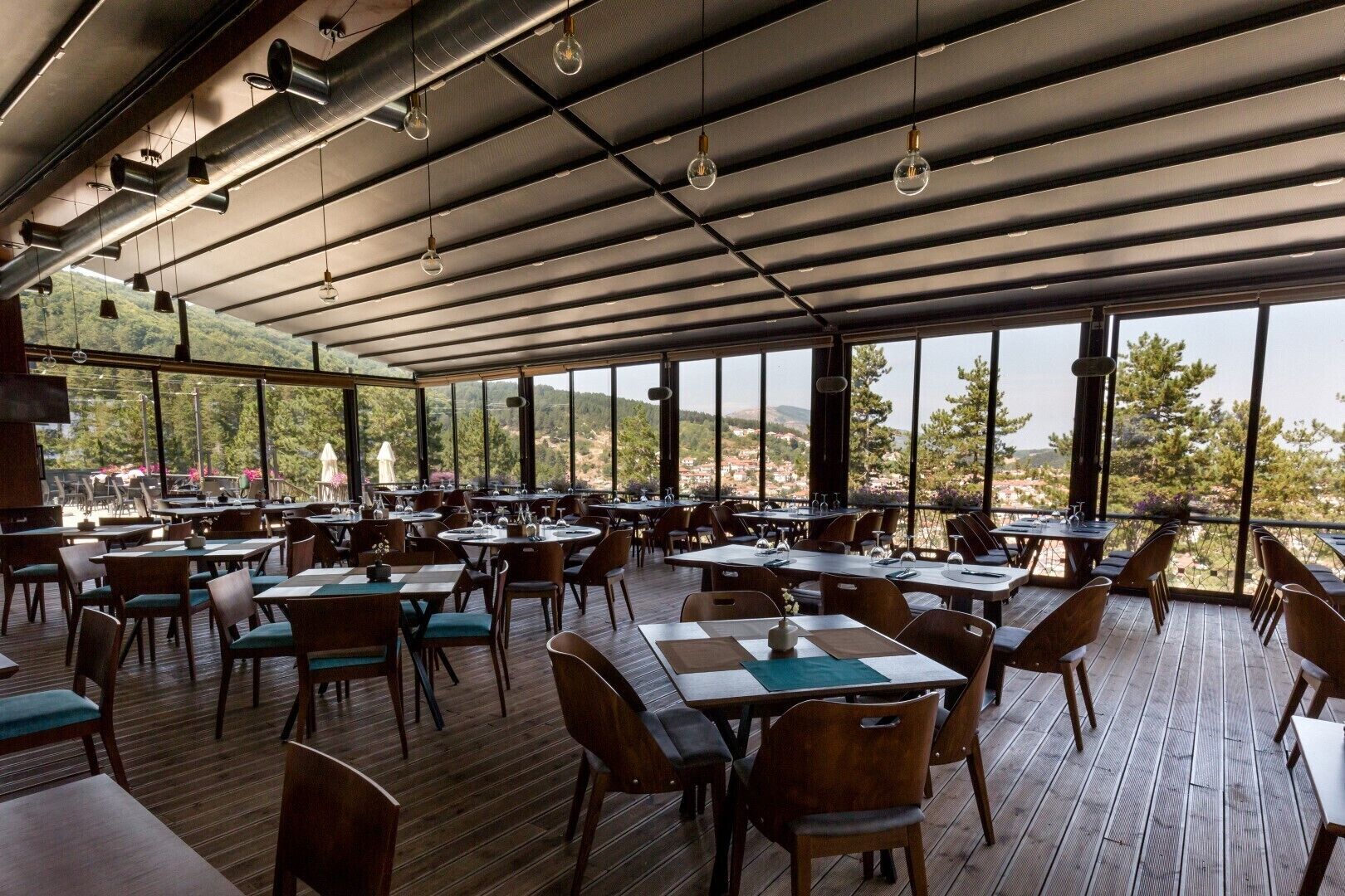 A spacious outdoor dining area with multiple tables and chairs, large windows offering scenic views, and hanging pendant lights.