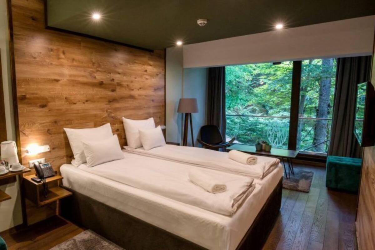 A modern bedroom with a large bed, a wooden accent wall, a bedside table with a lamp, and a view of the outdoors.