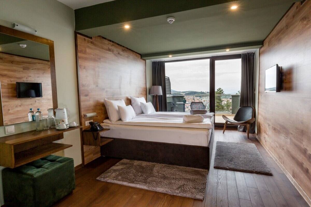 A modern hotel room with a large bed, a desk with a chair, a TV, and a city view.