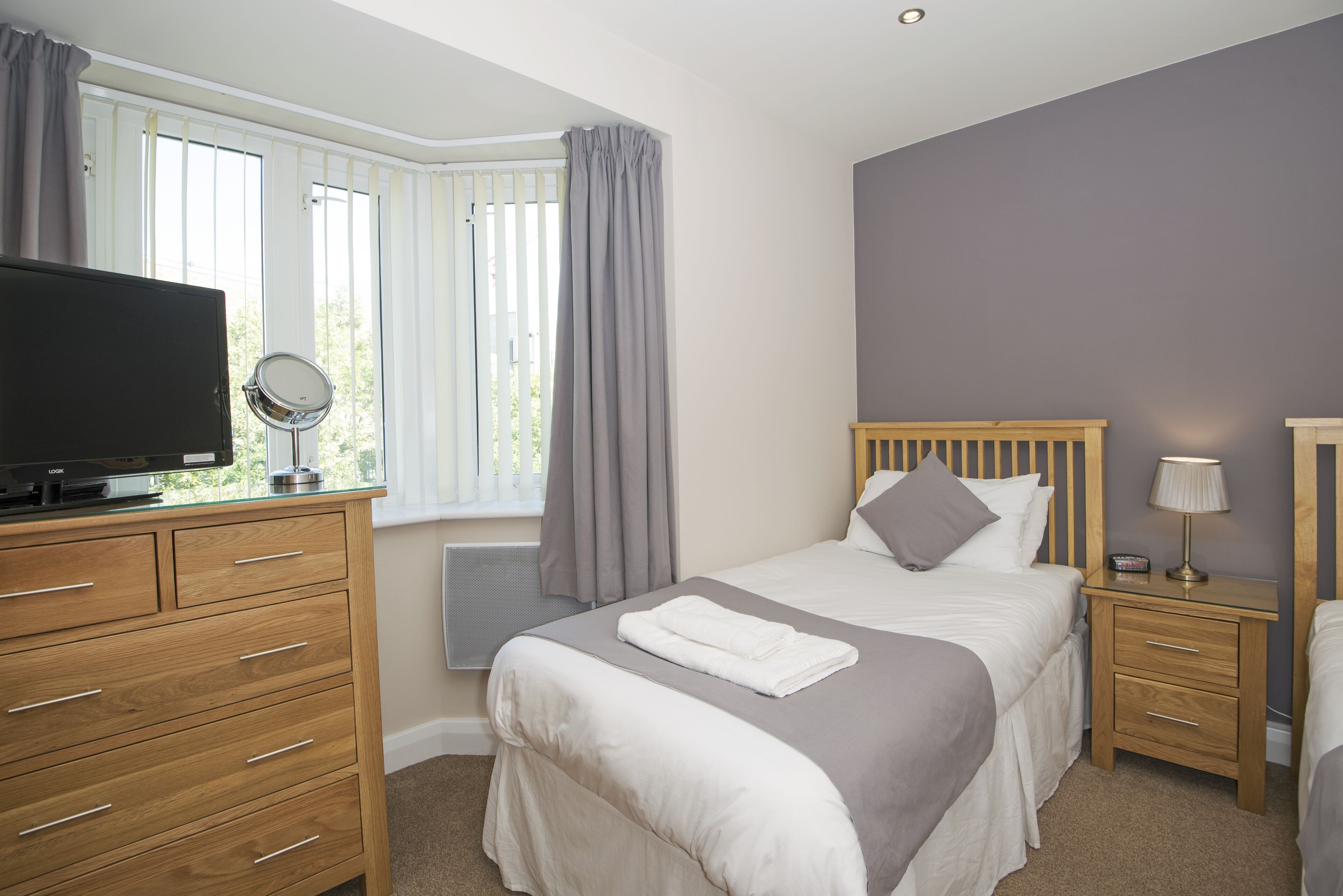 A bedroom with a bed, a TV on a wooden dresser, a bedside table with a lamp, and a window with curtains.