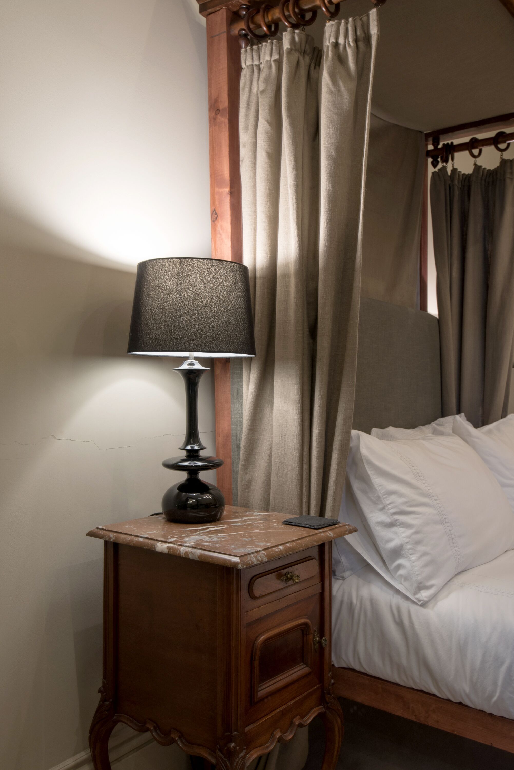 A wooden bedside table with a lamp, a bed with white linens, and a wooden headboard.