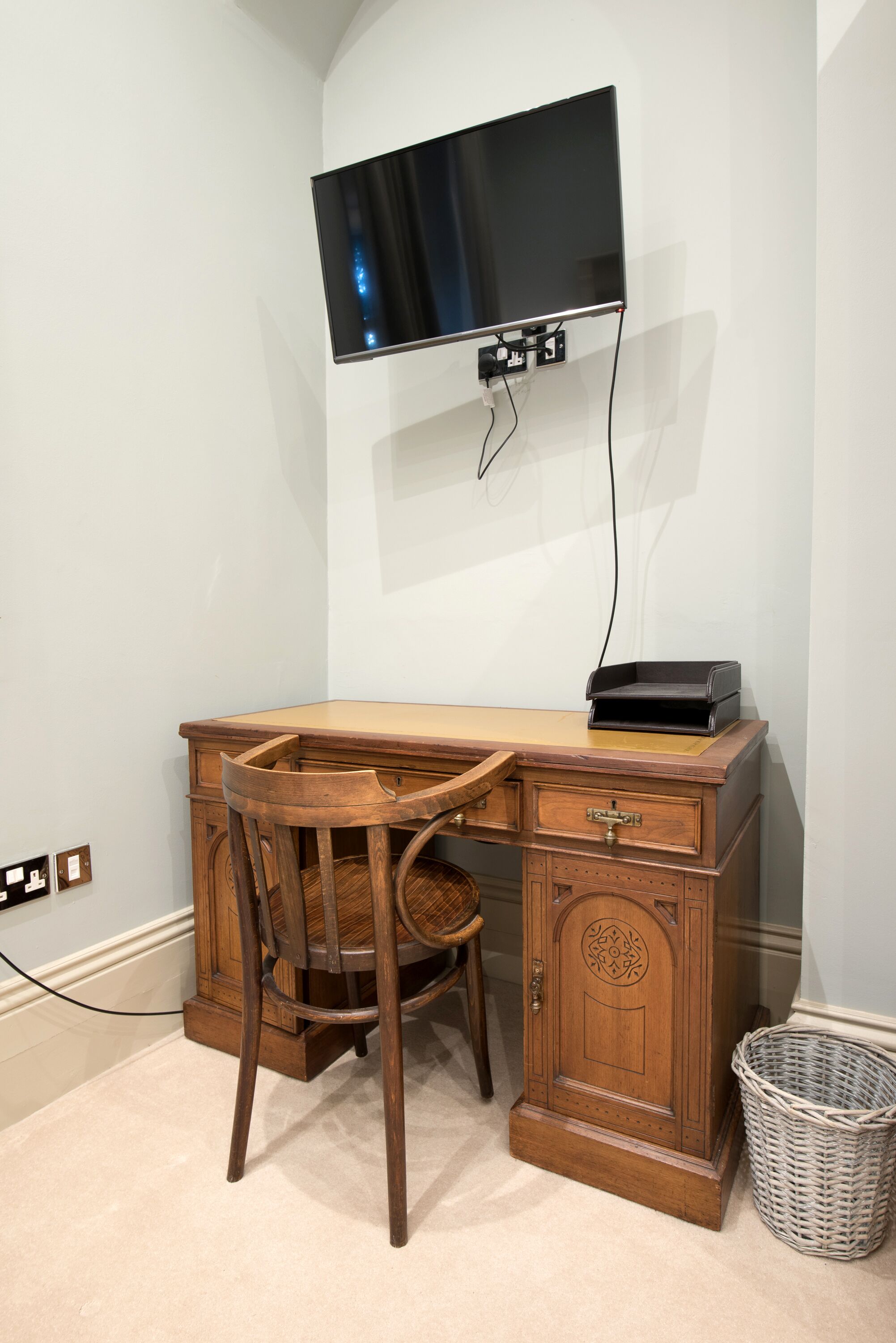 A corner desk with a chair, a flat-screen TV mounted on the wall, a basket, and a power outlet.