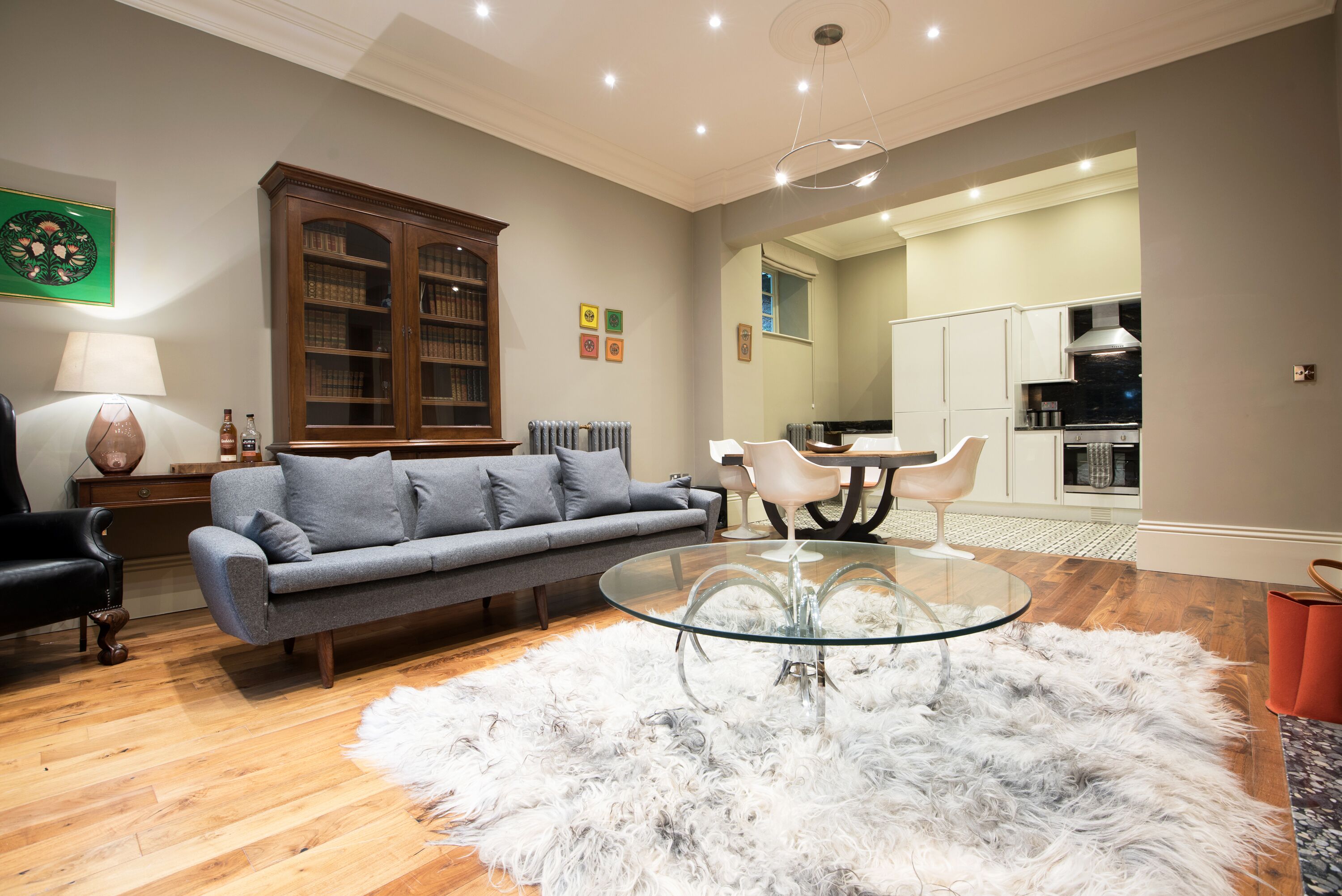 A living room with a grey sofa, a glass coffee table, and a white furry rug. There is a wooden cabinet with glass doors, a lamp, and a chair. The room has wooden flooring and a ceiling with recessed lighting.