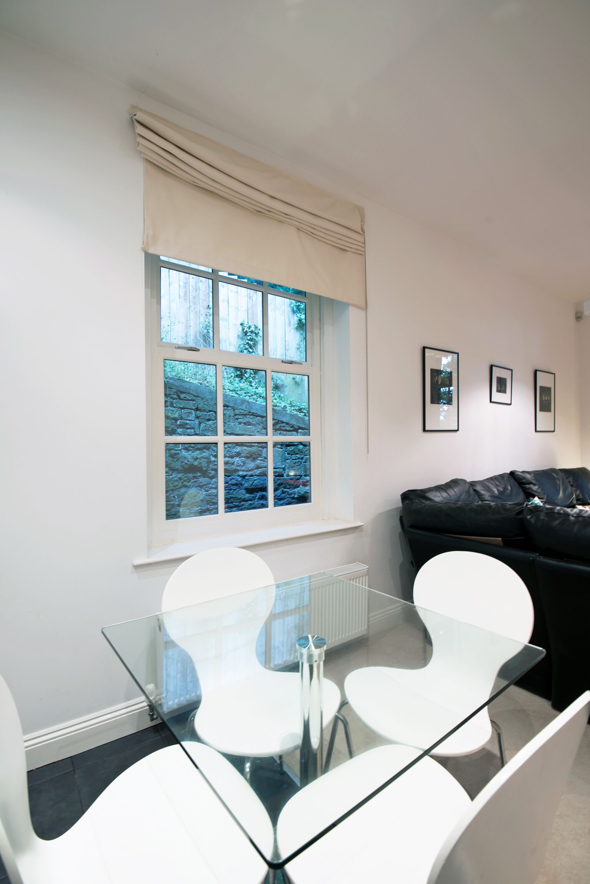 A modern room with a glass table and white chairs, a black leather sofa, and a window with a view of greenery.