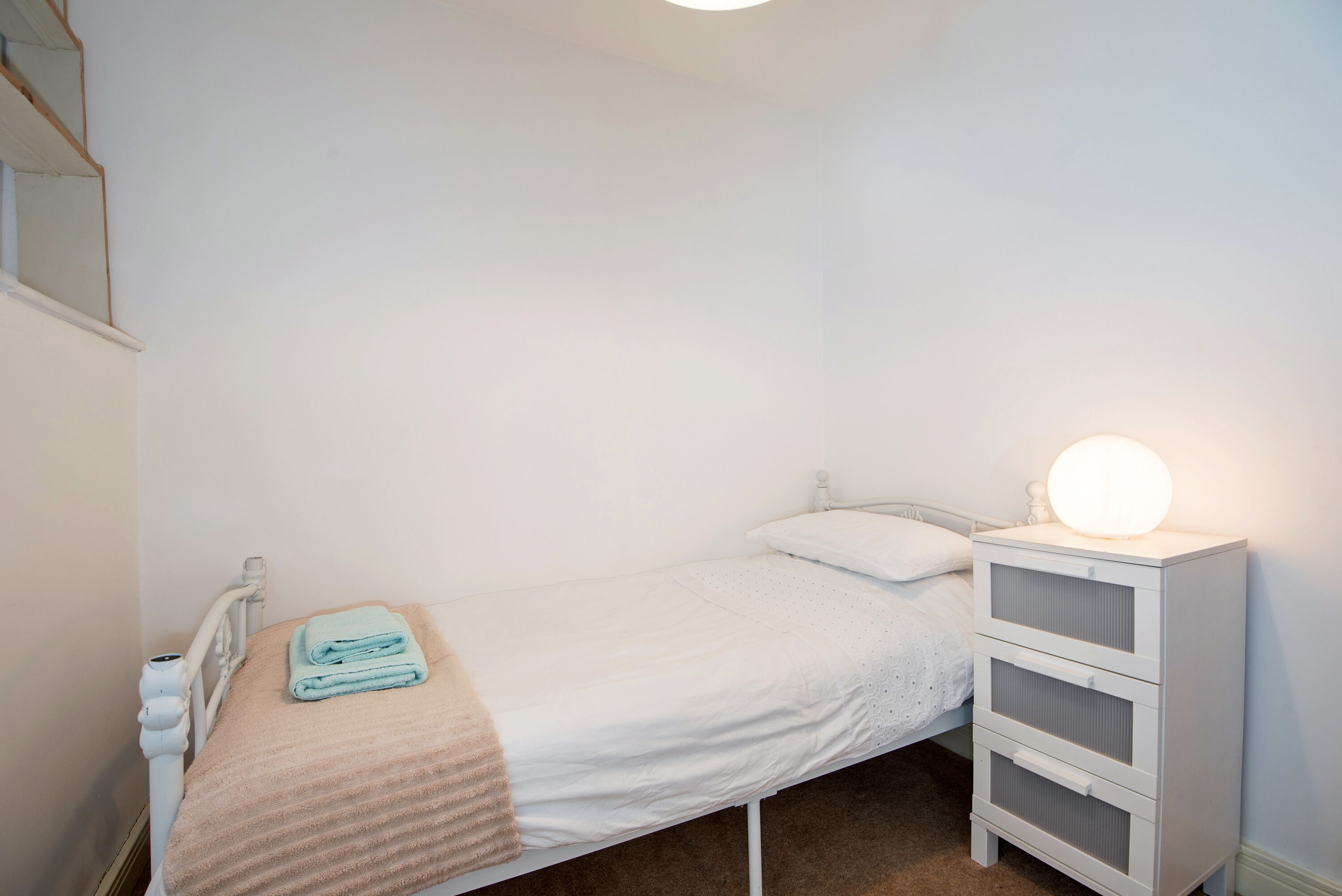 A small, single-bed room with a white bed, a bedside table with a lamp, and a white wardrobe.