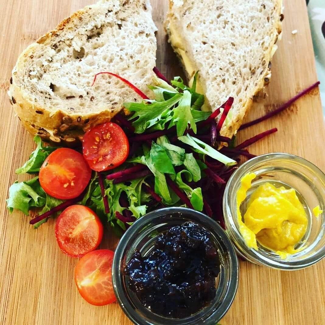 Un sándwich con ensalada, un cuenco de mermelada negra y un tazón pequeño de mostaza amarilla sobre una superficie de madera.