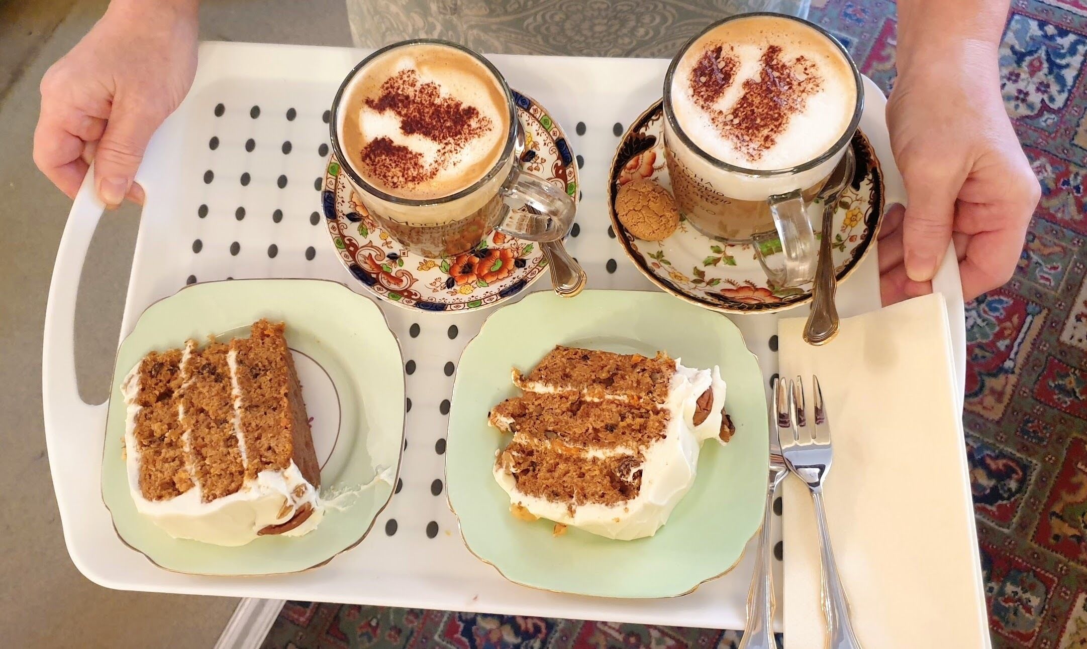 A tray with two plates of carrot cake and two cups of coffee with whipped cream and cocoa powder on a table with a polka dot tablecloth.
