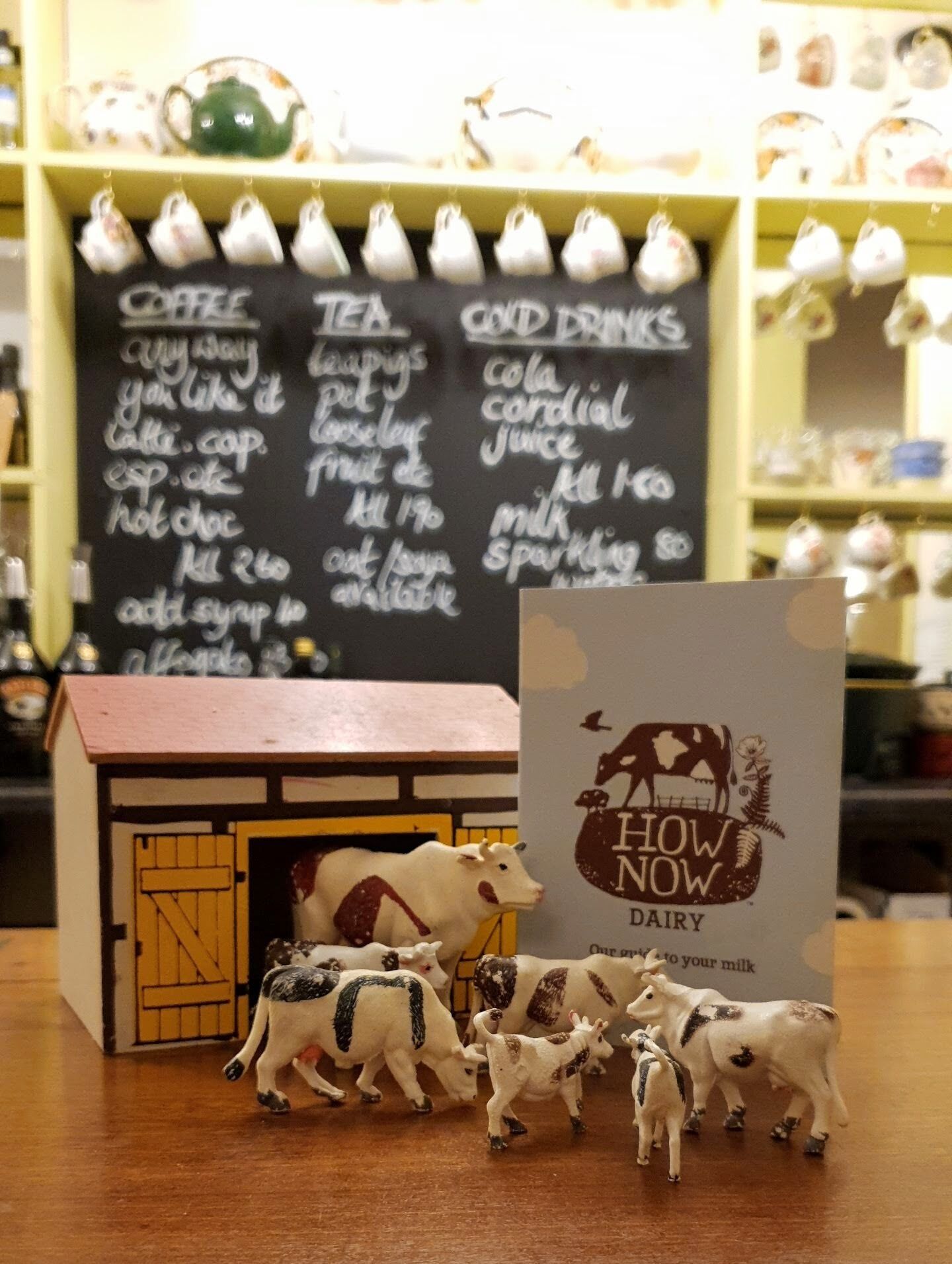 A small model of a barn with miniature cows in front of a chalkboard menu listing coffee, tea, and cold drinks.