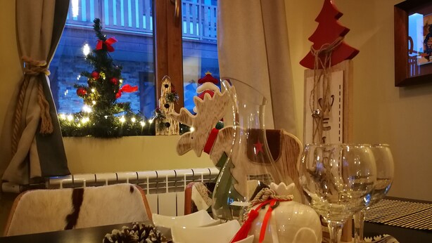 A dining table with a glassware set, a white bowl, and pine cones. A Christmas tree with lights is visible through a window.