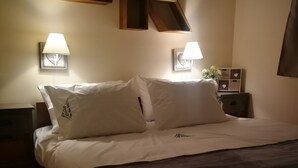 A neatly made bed with white linens and a dark brown throw blanket. There are two bedside lamps with white shades, a small bedside table with a decorative item, and a wall-mounted light fixture.