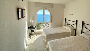 Ein Hotelzimmer mit zwei Betten, einem kleinen Tisch, einer Lampe und Meerblick durch ein großes Rundbogenfenster.