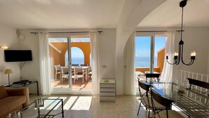 Ein geräumiges Wohnzimmer mit einer großen Glasschiebetür, die auf einen Balkon führt, einem Essbereich mit einem Glastisch und einem an der Wand befestigten Fernseher.