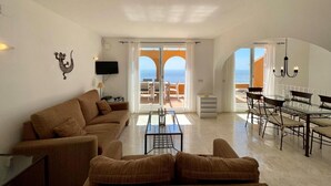 Ein geräumiges Wohnzimmer mit einer Couch, einem Couchtisch und einem Fernseher. Es gibt einen Balkon mit Tisch und Stühlen.