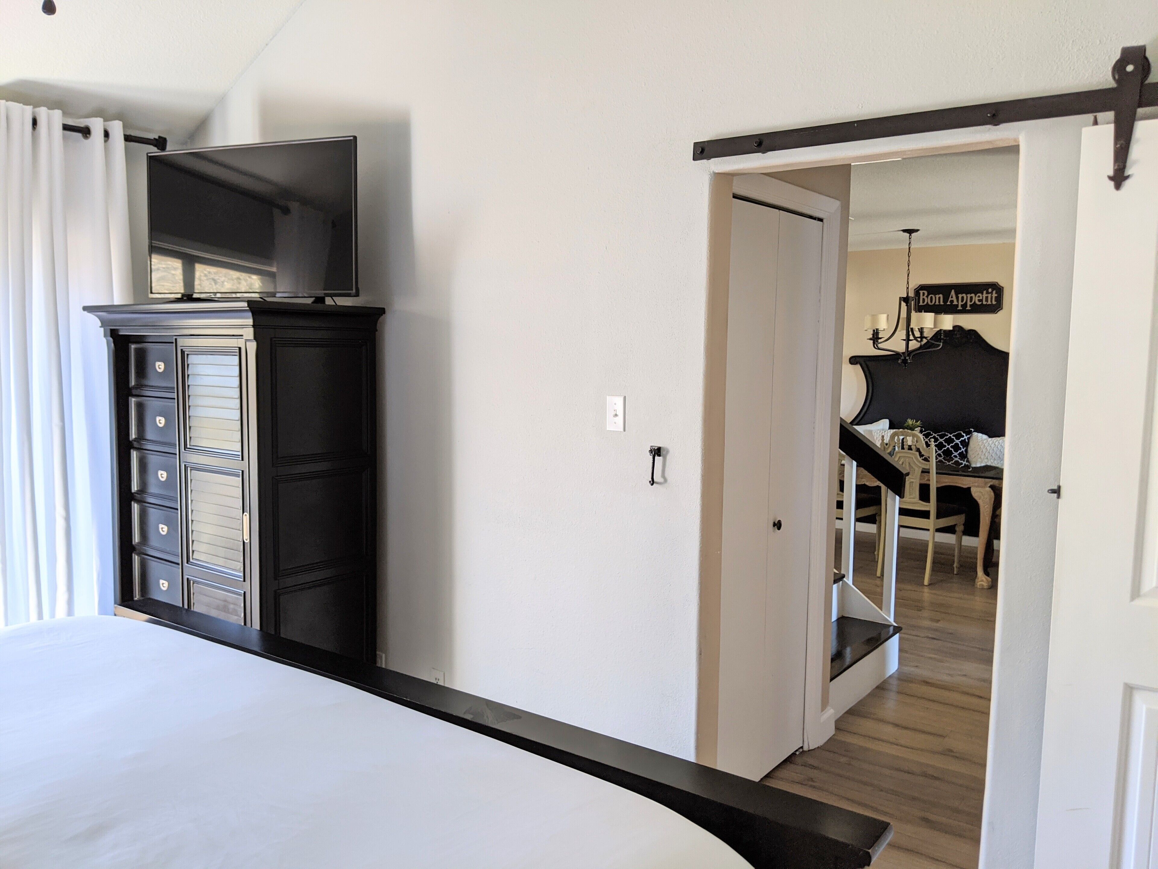 Een slaapkamer met een bed, een dressoir en een televisie erboven. Er is een deur die leidt naar een eetgedeelte met een tafel en stoelen.