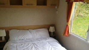 A single bed with white bedding, two bedside lamps, wooden headboard, and a window with curtains. - Steading Holidays - Croft Caravan (Acharacle)