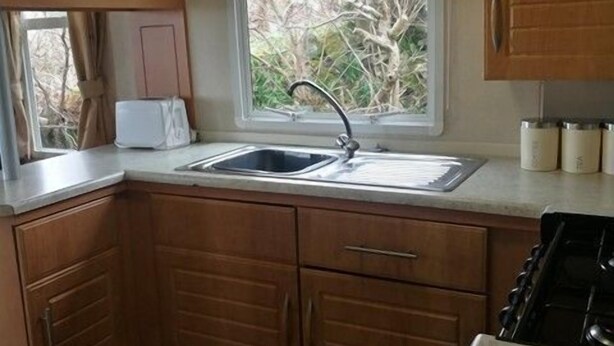 A compact kitchen with wooden cabinets, a window, a sink, and a stove. - Steading Holidays - Croft Caravan (Acharacle)