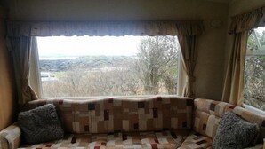 A cozy living room with a quilted sofa facing a window with a view of a hilly landscape. - Steading Holidays - Croft Caravan (Acharacle)