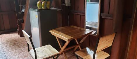 Una habitación compacta con una pequeña mesa de madera y dos sillas, un refrigerador con alimentos, y una ventana que ofrece vistas al exterior.