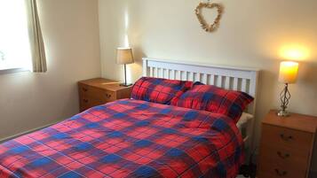 A bedroom with a bed, two bedside tables, a lamp, a heart-shaped decoration on the wall, and a window with curtains.