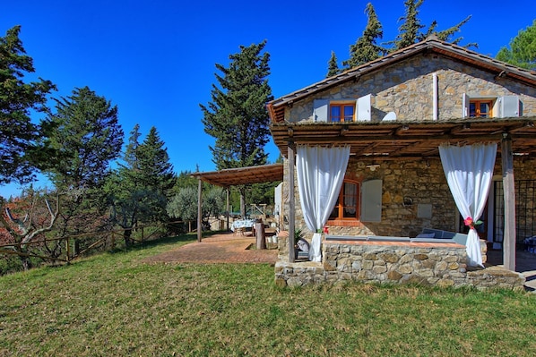 A stone house with a covered porch, a dining area with outdoor seating, and a view of surrounding trees.