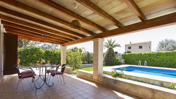 Un patio techado con piscina, mobiliario exterior y vistas a la vegetación y edificios circundantes.