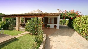 Una casa con porche cubierto, camino de piedra y jardín con diversas plantas y flores.