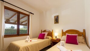 Habitación de hotel con dos camas, cada una con cabecero de madera, ropa de cama color beige y almohada rosa. Hay una ventana con cortinas, una mesita de noche con lámpara y un cuadro en la pared.