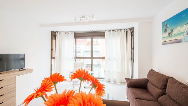 A modern living room with a brown sofa, a TV on a wooden stand, and a large window with curtains. - "The QUEEN" New, fancy and relaxing. Beach & WIFI (València)