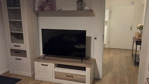 Un salon avec un téléviseur à écran plat posé sur un meuble télé en bois, une armoire blanche avec des tiroirs, une petite table et une porte donnant sur une autre pièce.
