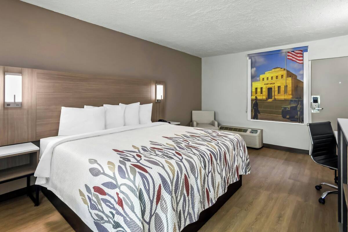 A hotel room with a large bed, a desk, a chair, a window with a view of a building, and a wall-mounted air conditioner.