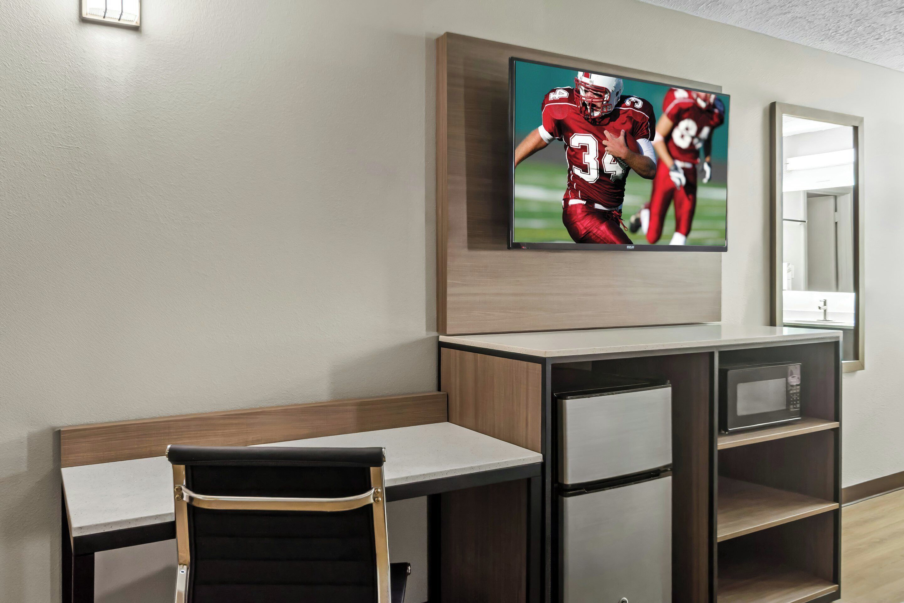 A modern hotel room with a flat-screen TV mounted above a wooden desk and chair, a microwave on a shelf, and a mirror on the wall.