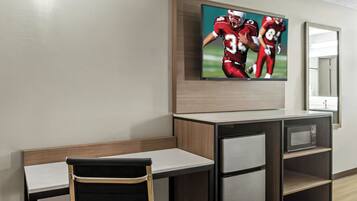A modern hotel room with a flat-screen TV mounted above a wooden desk and chair, a microwave on a shelf, and a mirror on the wall.