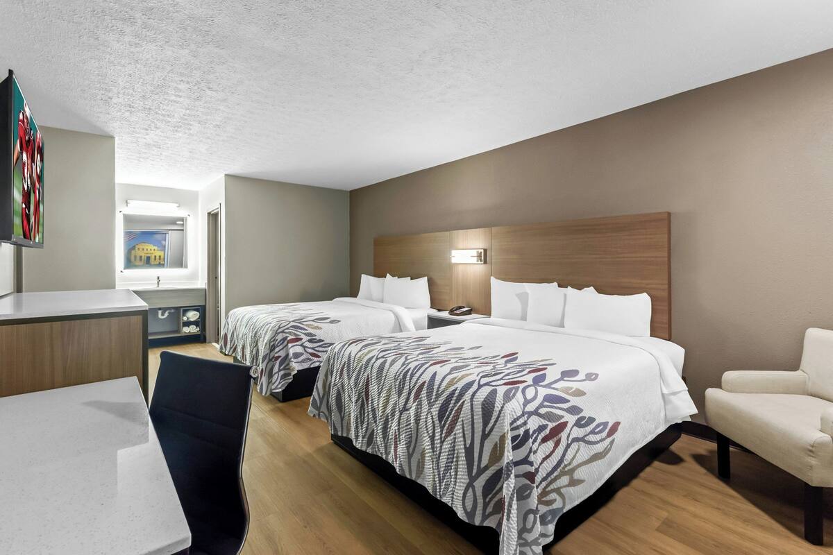 A hotel room with two beds, a desk, a chair, and a view of a building through a window.