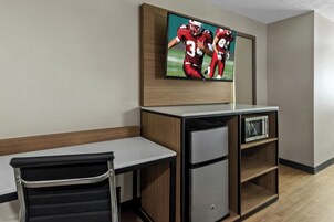 Habitación compacta con escritorio, una silla, televisor montado en la pared y un refrigerador pequeño.