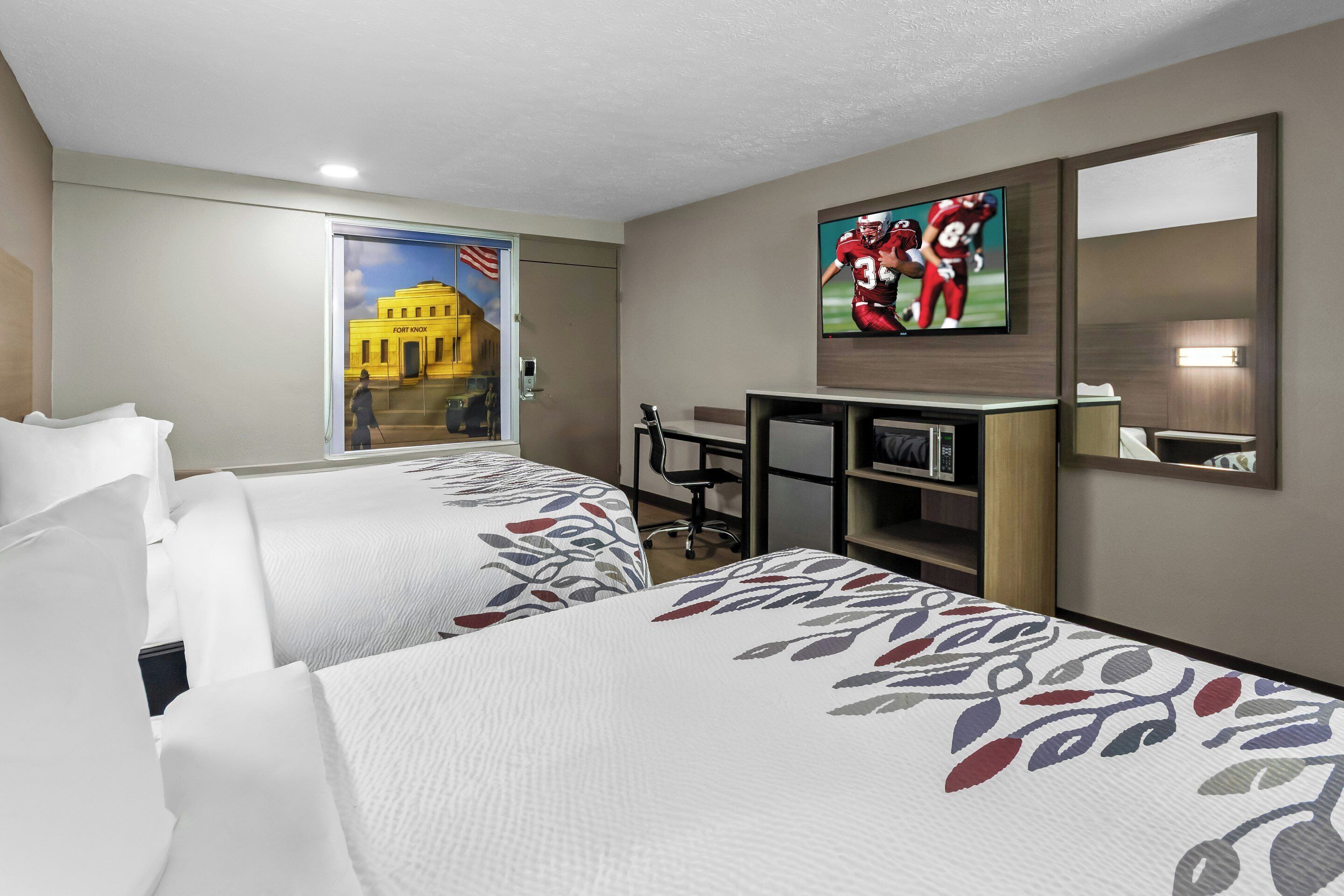 A hotel room with two beds, a desk, a TV, and a window with a view of a building.