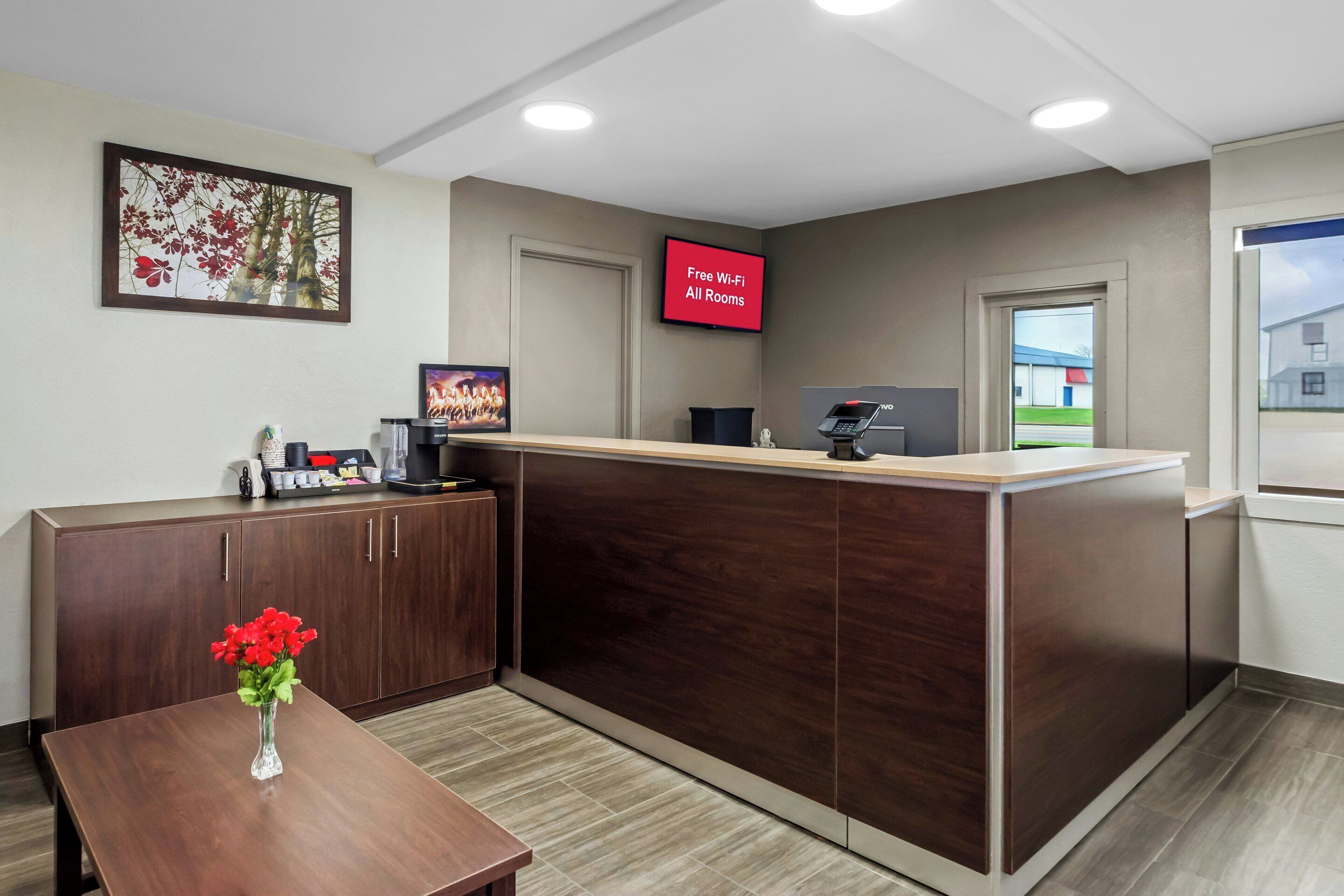 A reception desk with a digital screen, a wooden cabinet, a vase with red flowers, and a framed picture on the wall.